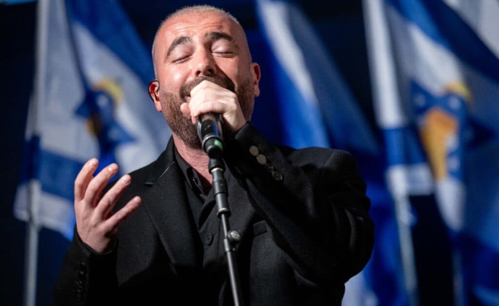 Omer Adam sings into a microphone on stage with Israeli flags in the background.