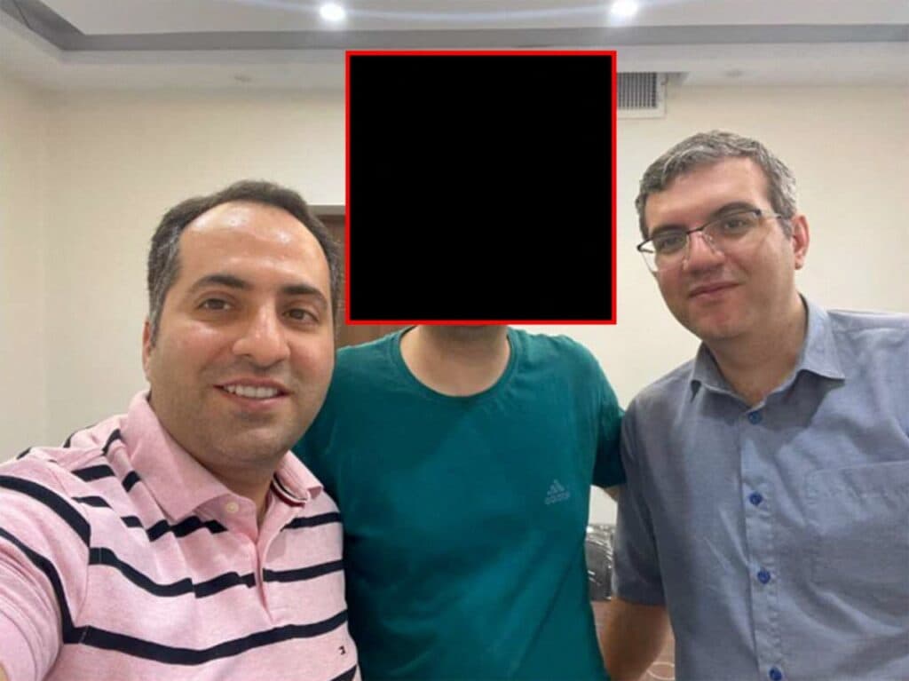 Three men standing indoors, with the face of the man in the center obscured.