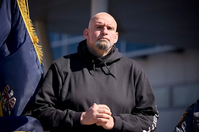 John Fetterman stands outdoors while listening to speakers.