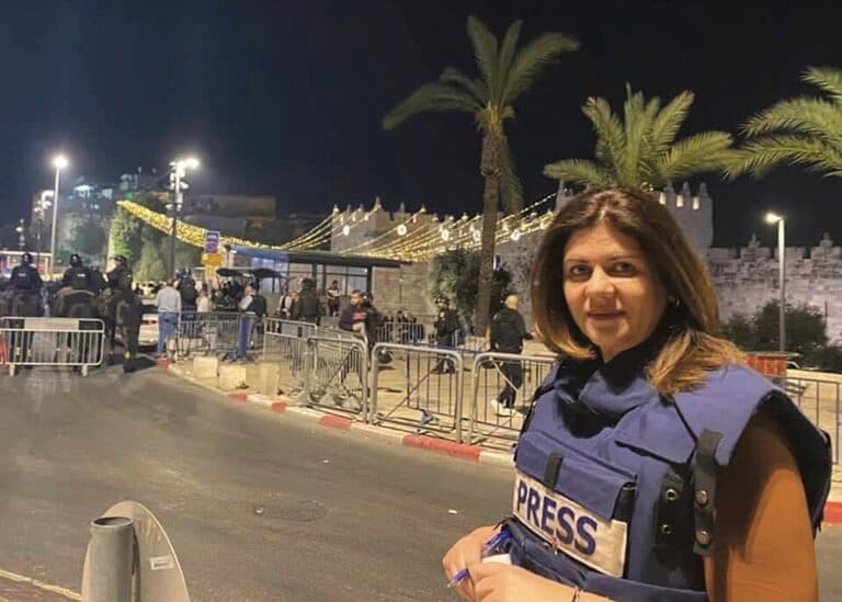 Shireen Abu Akleh stands wearing a press vest near a street scene at night.