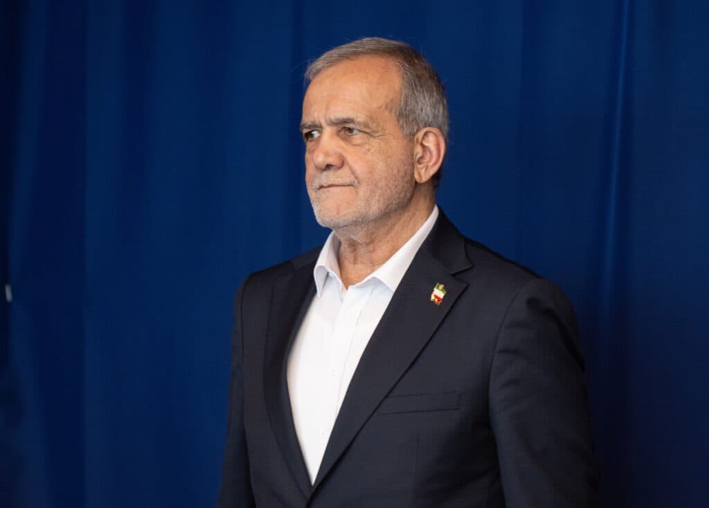 Iranian President Masoud Pezeshkian standing in front of a blue curtain.