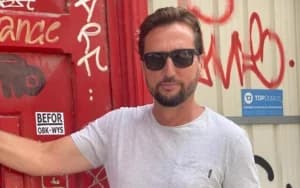 Portrait of Haim Kalomiti wearing sunglasses and a gray T-shirt, standing in front of a red wall with graffiti.