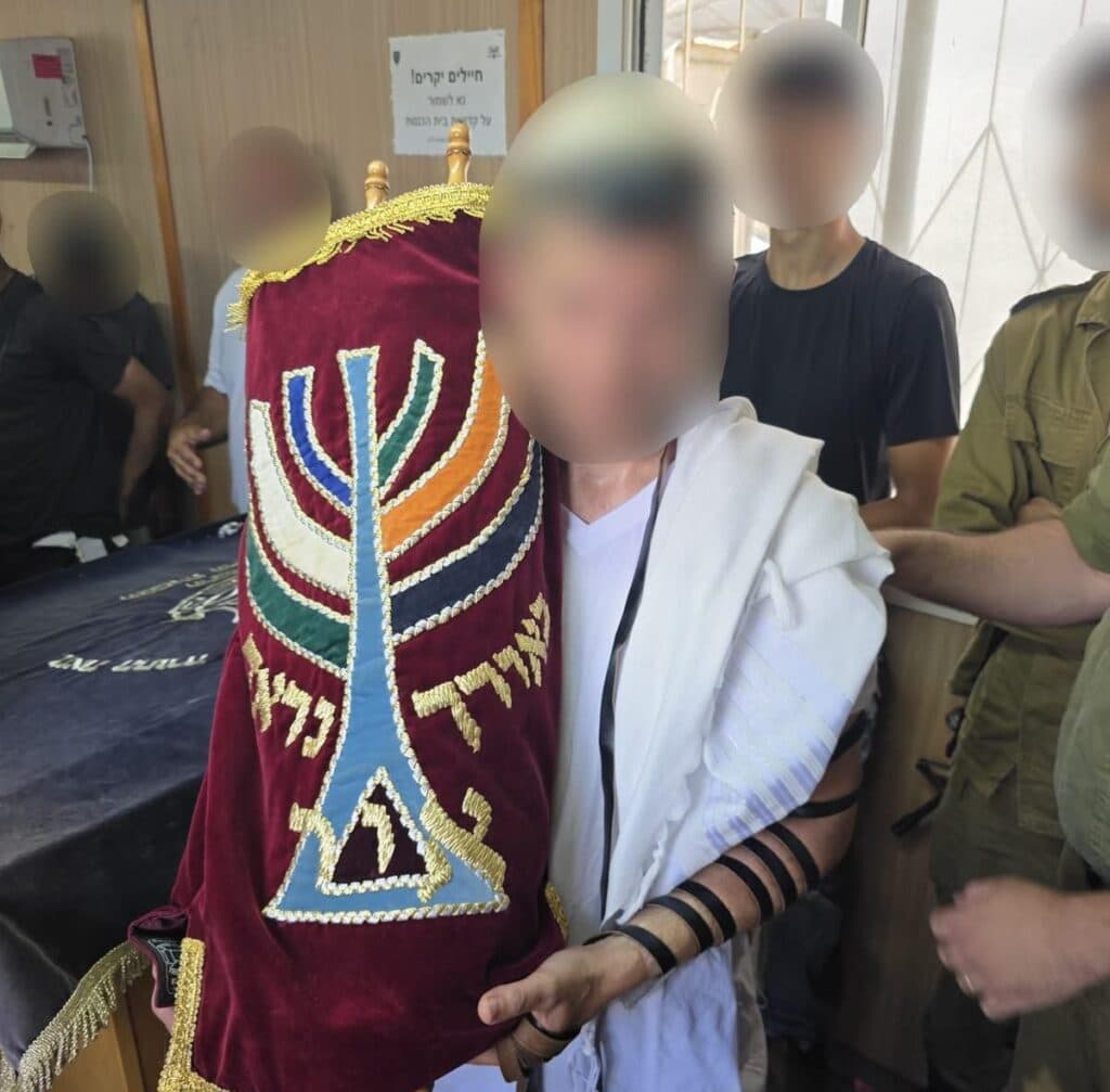 An Israeli reservist wearing tefillin and holding a Torah scroll during a bar mitzvah ceremony with fellow soldiers.