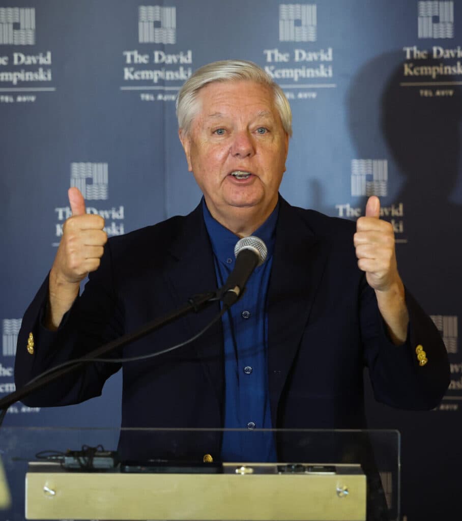 U.S. Senator Lindsey Graham speaks at a podium in Tel Aviv with Israeli and American flags beside him.
