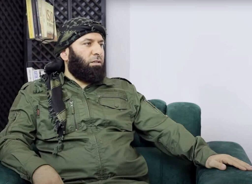 A man in military-style clothing sits on a sofa indoors.