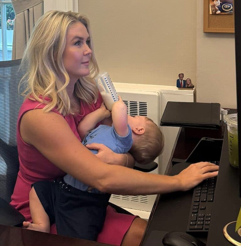 Karoline Leavitt sits at a desk working on a laptop while feeding her baby with a bottle.
