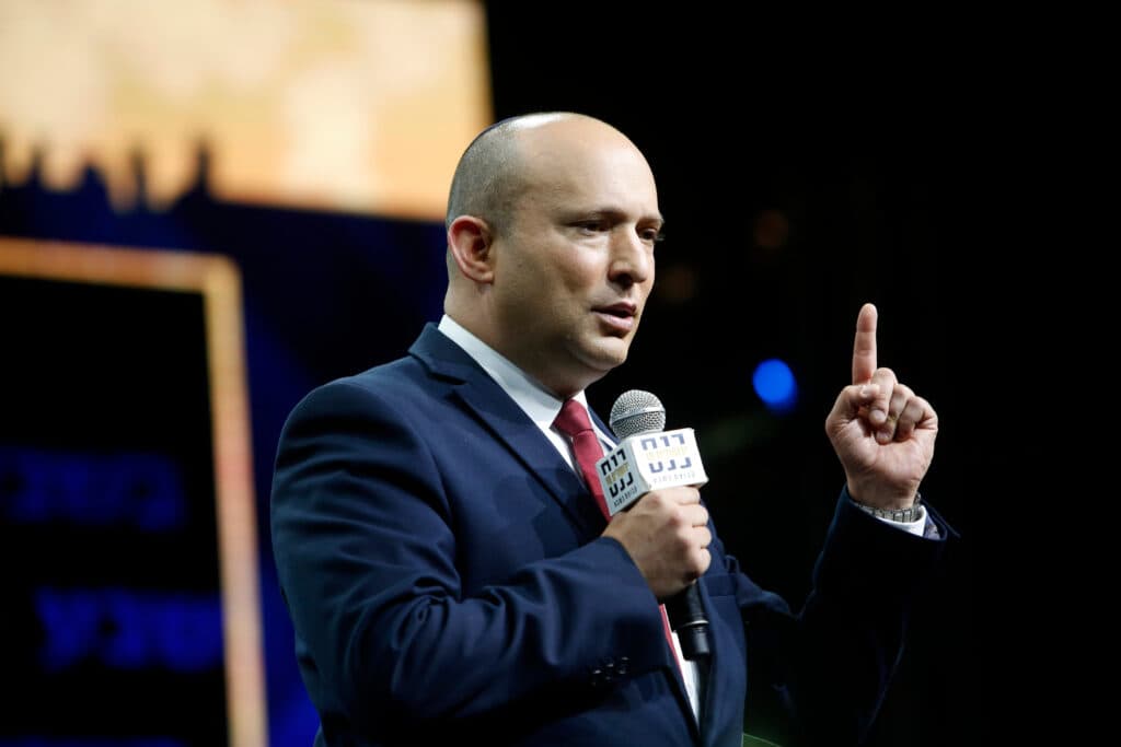 Naftali Bennett speaking into a microphone while raising one finger.