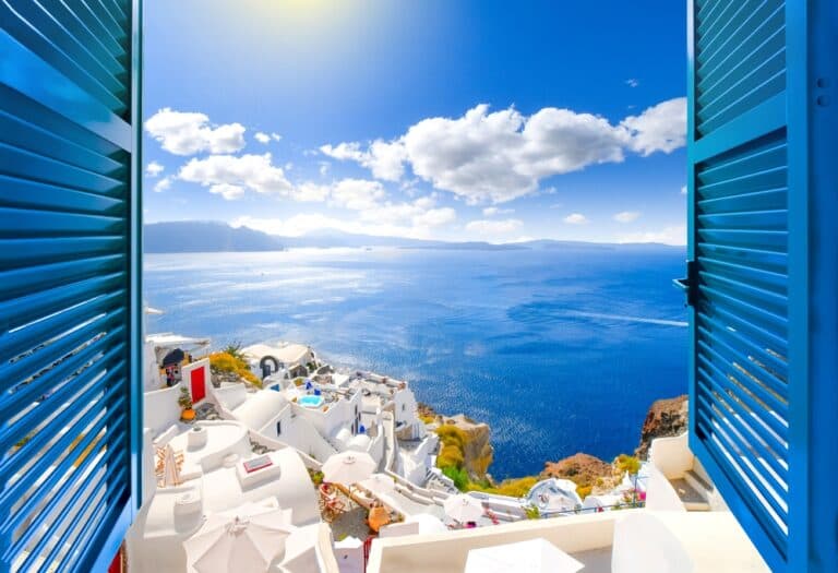 Hillside view through an open window with blue shutters of the caldera, sea and white village of Oia on the island of Santorini, Greece.