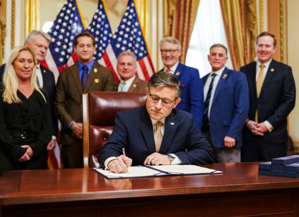 A U.S. lawmaker signs legislation at a desk while members of Congress stand behind him with American flags.